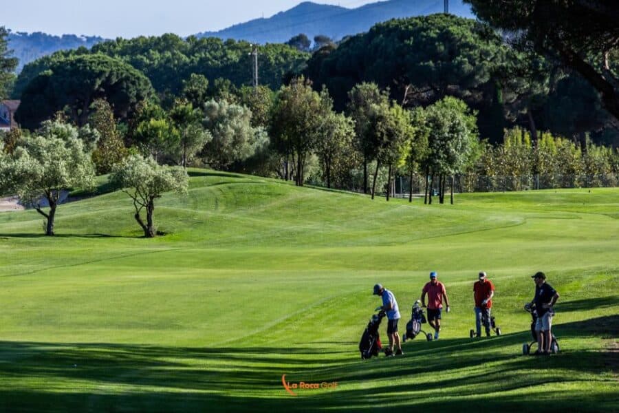 Grupo de cuatro jugadores amateurs recorriendo un hoyo en La Roca Golf, rodeados de olivos centenarios y pinos mediterráneos. El campo verde esmeralda ondulado se extiende hacia las montañas del fondo, creando un paisaje natural que ejemplifica la perfecta integración del campo con el entorno. La luz dorada del atardecer realza la experiencia social y deportiva que caracteriza al golf amateur.