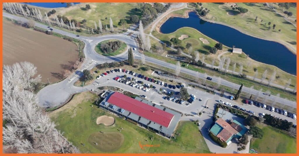 Vista aérea de las instalaciones principales de La Roca Golf, mostrando la Casa Club con techo rojo característica, putting green de prácticas, amplias zonas de aparcamiento y acceso directo al campo con lago. Ubicación privilegiada en Barcelona con excelente conectividad