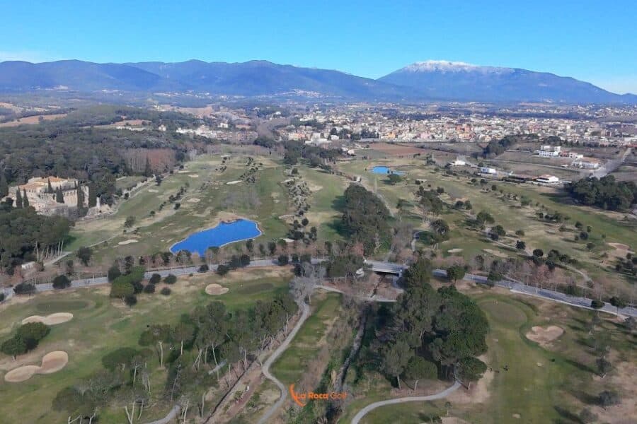 Vista aérea del campo de golf La Roca Golf en Barcelona, mostrando sus 18 hoyos, lagos artificiales y el castillo histórico de La Roca, rodeado de bosque mediterráneo con el Montseny nevado al fondo