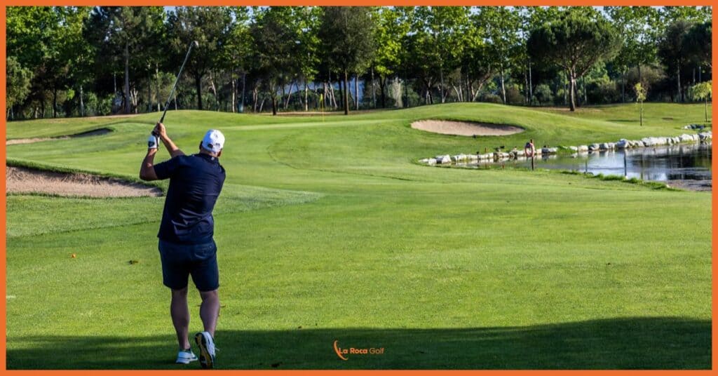 Jugador ejecutando un approach hacia un green protegido por agua y bunkers en La Roca Golf. La imagen captura la esencia de nuestro diseño estratégico, donde cada hoyo presenta un reto equilibrado entre riesgo y recompensa. Desde esta posición, el jugador debe evaluar cuidadosamente su golpe, considerando el lago que bordea el green con su característica orilla de piedra, y el bunker estratégicamente ubicado. El entorno natural, con sus pinos mediterráneos cuidadosamente podados, enmarca la escena y proporciona una referencia visual clara para la estrategia de juego. La calidad del césped y el mantenimiento impecable del campo reflejan nuestro compromiso con la excelencia en cada detalle.