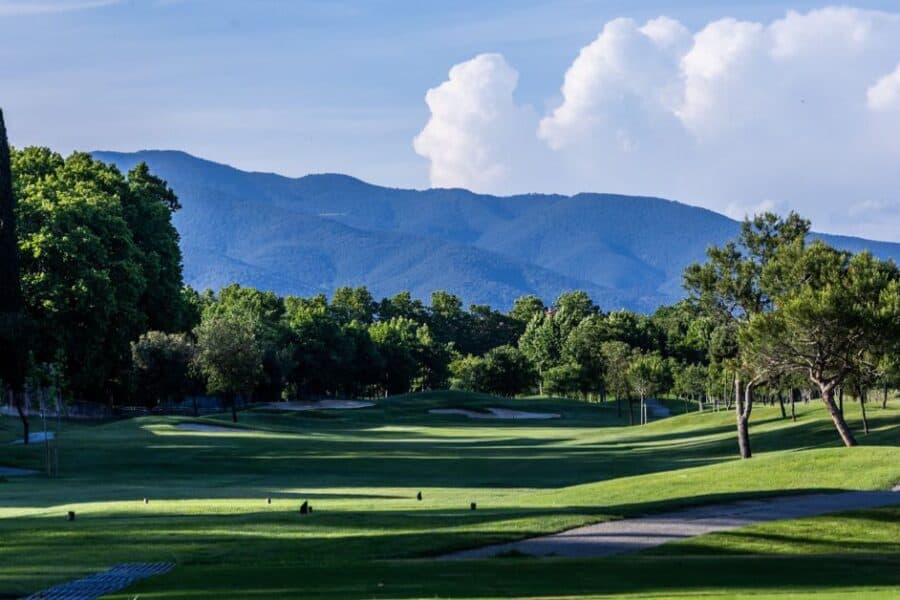 Scenic golf course with manicured fairway, surrounded by lush trees and blue mountains in the background under a bright sky with fluffy clouds