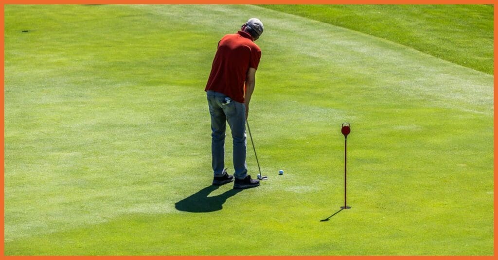 La imagen muestra a un golfista en el putting green de La Roca Golf, concentrado en ejecutar un putt. Lleva una camiseta roja y jeans, y sostiene un putter mientras se prepara para golpear la bola blanca hacia el hoyo marcado con una bandera. El césped del green luce impecablemente cuidado, con diferentes tonalidades de verde que indican los sutiles desniveles característicos de los greens de calidad. Esta imagen ilustra perfectamente el área de putting de 1.000 m² que ofrece La Roca Golf, donde los jugadores pueden practicar todo tipo de putts con diferentes caídas y distancias, elementos fundamentales para mejorar su juego corto y reducir el número de golpes en cada recorrido.