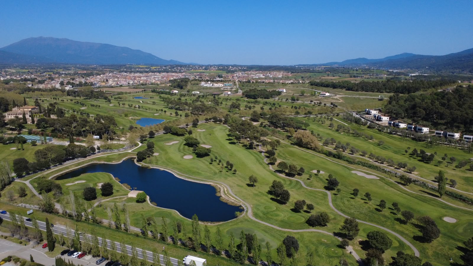 Vista aérea de La Roca Golf con sus lagos y la carretera de acceso, el pueblo del Vallès Oriental y la sierra del Montseny al fondo