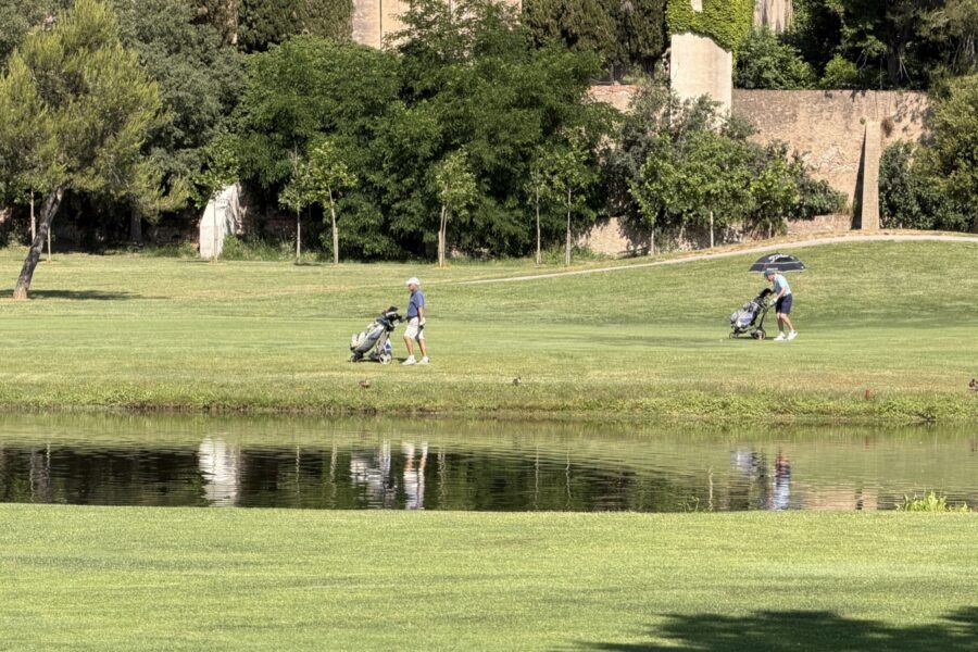 Dos jugadores caminando con sus carritos manuales por una calle llana de La Roca Golf, junto a uno de los lagos del campo.