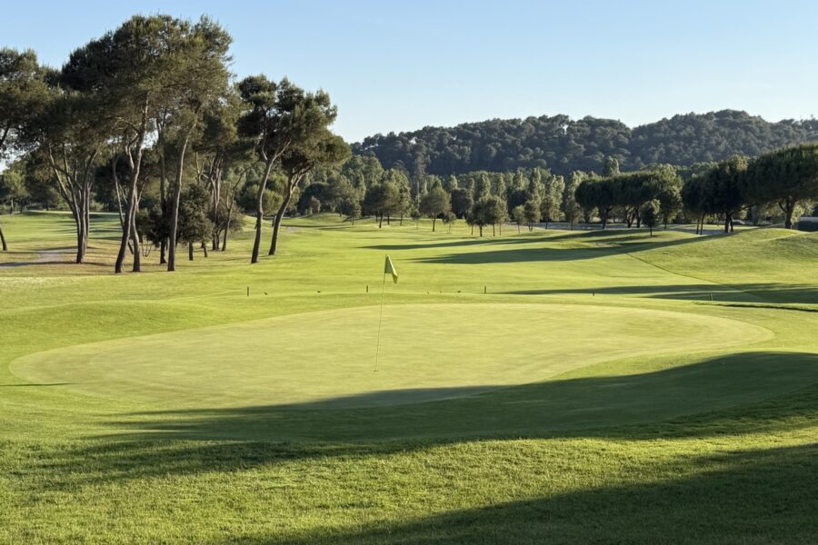 Green del hoyo 1 de La Roca Golf entre pinos, con luz baja y sombras largas sobre la calle